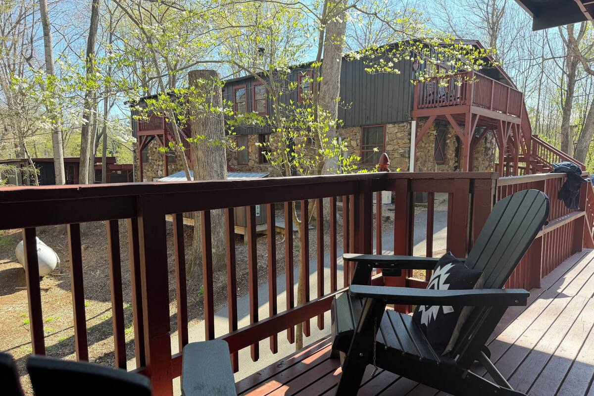 A wooden deck with chairs and a table, surrounded by trees and a stone building.