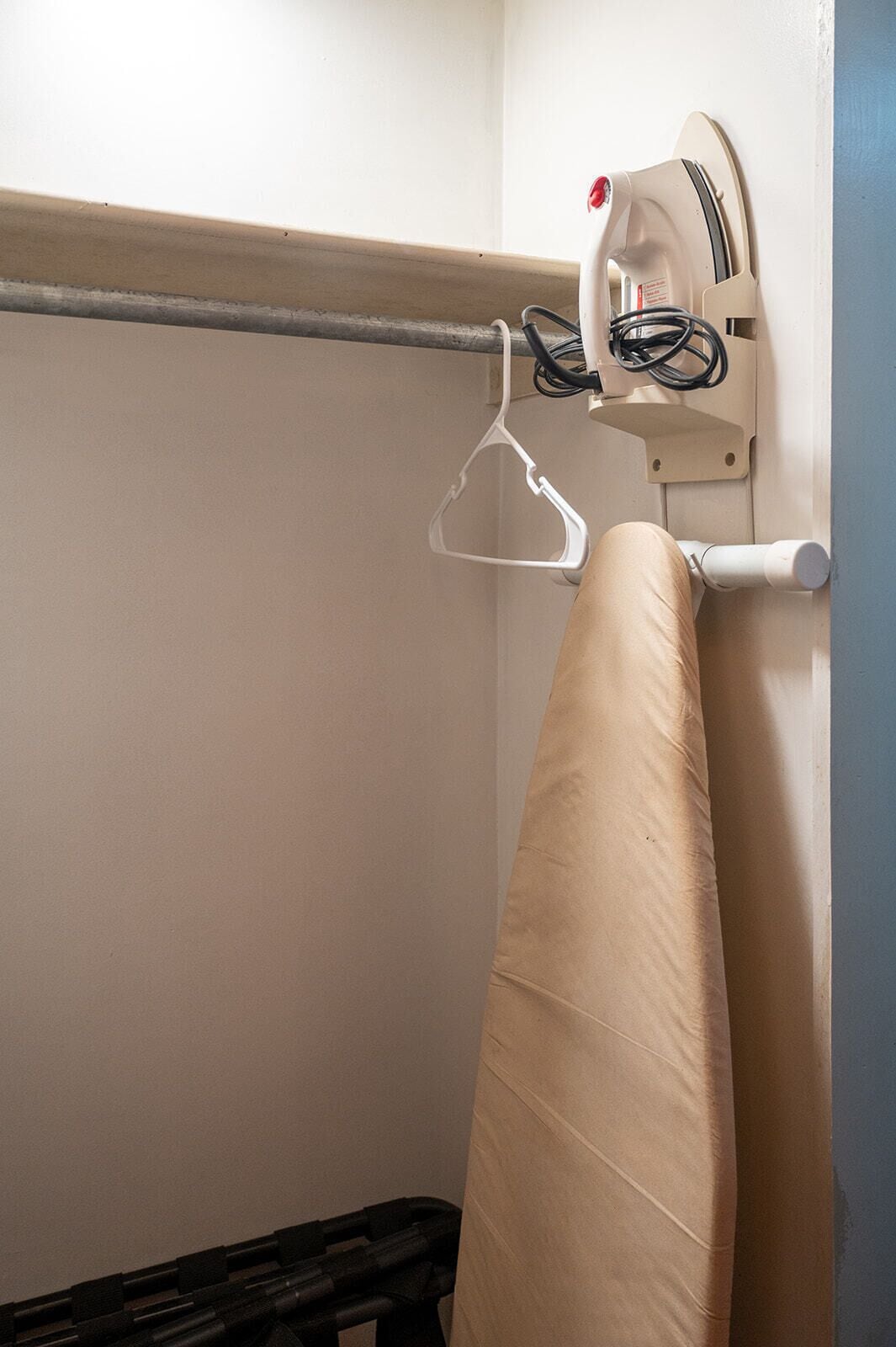 Un rincón de una habitación con una tabla de planchar fijada a la pared, un perchero y un cubo de basura negro.
