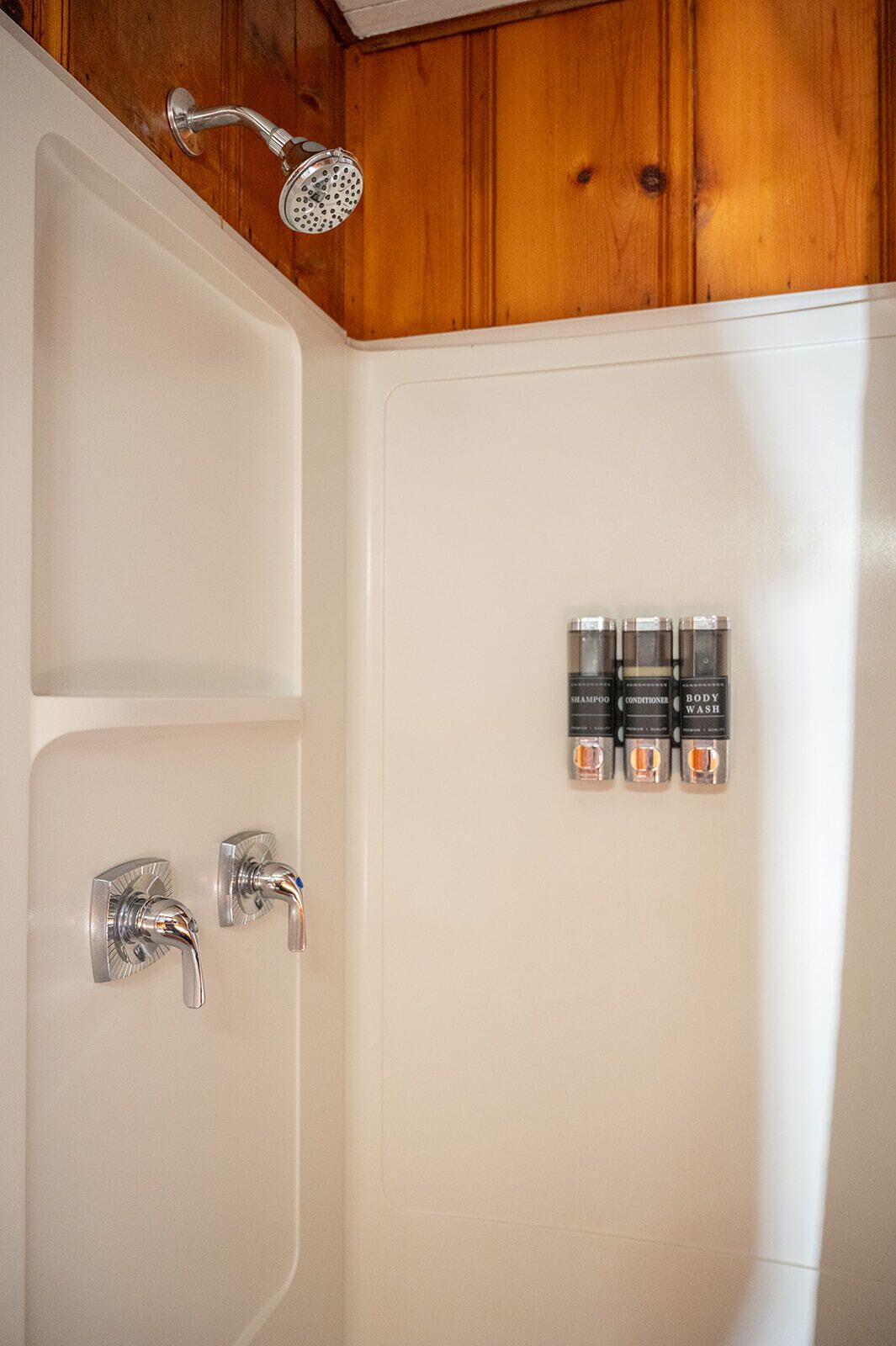 A bathroom with a shower, two faucets, and three bottles of shampoo, conditioner, and body wash.