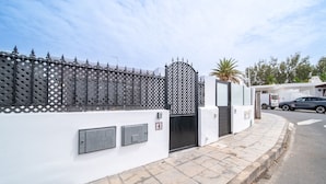 A gated residential area with a white wall, a metal gate, and a palm tree. - Casa Apolo (Puerto del Carmen)