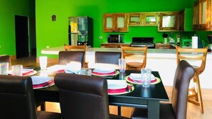 On aperçoit un coin repas avec une table dressée pour quatre personnes, entourée de chaises. Au fond, on distingue une cuisine aux murs verts et aux armoires en bois.