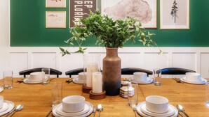 A dining table set with white plates, bowls, and cutlery, surrounded by chairs, with a vase of green foliage in the center.