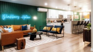 A modern living room with a green accent wall, a neon sign, a sofa, a coffee table, and a dining area with bar stools.