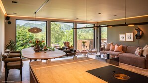 A modern living room with a wooden coffee table, a sofa, and a dining area with chairs. Large windows offer a view of greenery and hills.