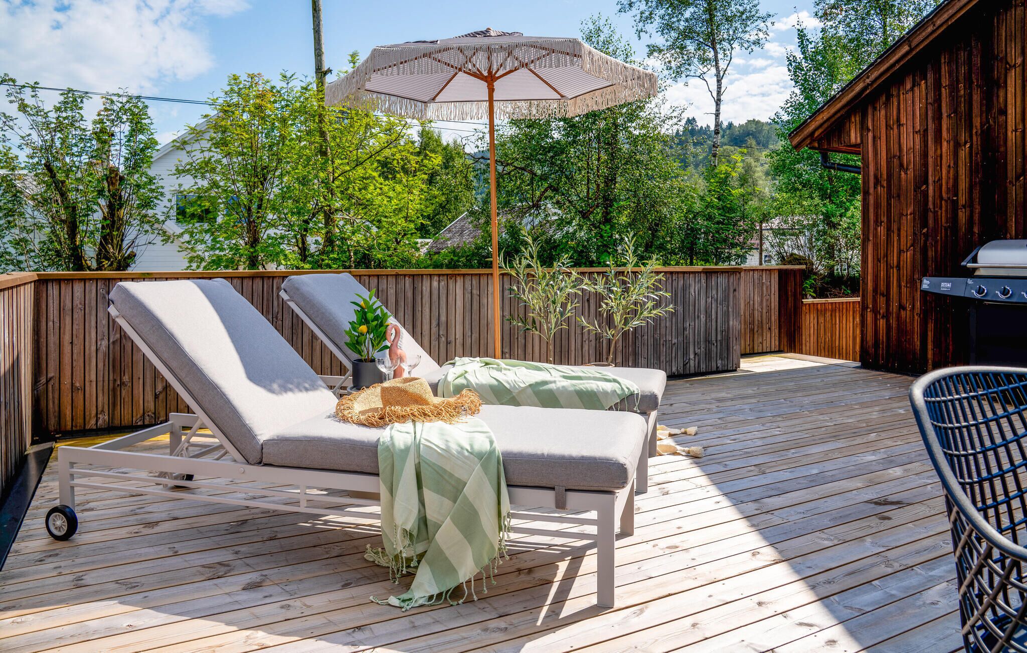 Une terrasse en bois avec deux chaises longues, une pergola dotée d’un auvent et un barbecue.
