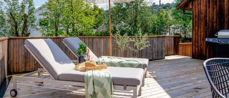 Une terrasse en bois avec deux chaises longues, une pergola dotée d’un auvent et un barbecue.