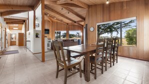 Un vaste espace dédié à la salle à manger, avec une table et des chaises en bois, de grandes fenêtres offrant une vue sur l’extérieur, et un coin cuisine à l’arrière-plan.