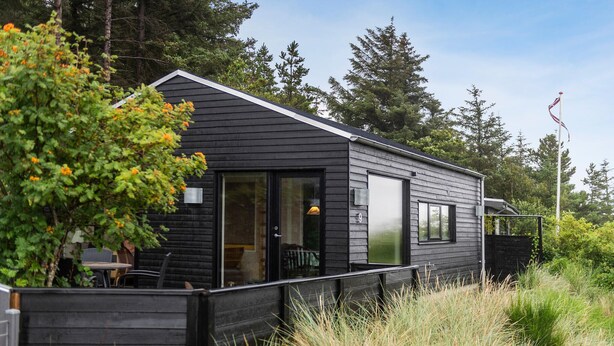 A modern cabin with a large sliding door entrance, a deck area with outdoor furniture, and a flagpole in the background. - 1 bedroom stunning home in Esbjerg V (Esbjerg V)