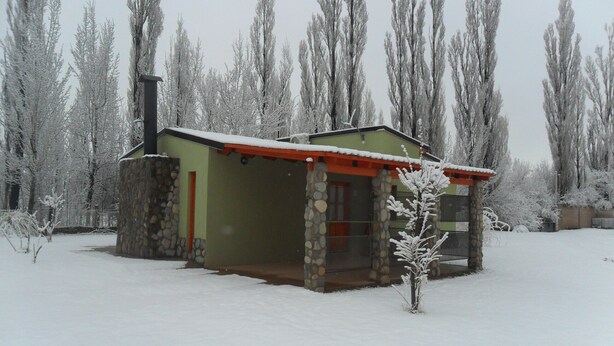 Ein mit Schnee bedecktes Blockhaus mit Steinaußenwand und rotem Dach, umgeben von hohen, kahl stehenden Bäumen.