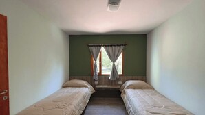 Ein kleines Zimmer mit zwei Einzelbetten, einem Fenster mit Vorhängen und einer Tür links.