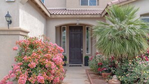 Une maison avec une porte d’entrée sombre, une façade de couleur claire et un petit jardin agrémenté de fleurs roses et d’un palmier.