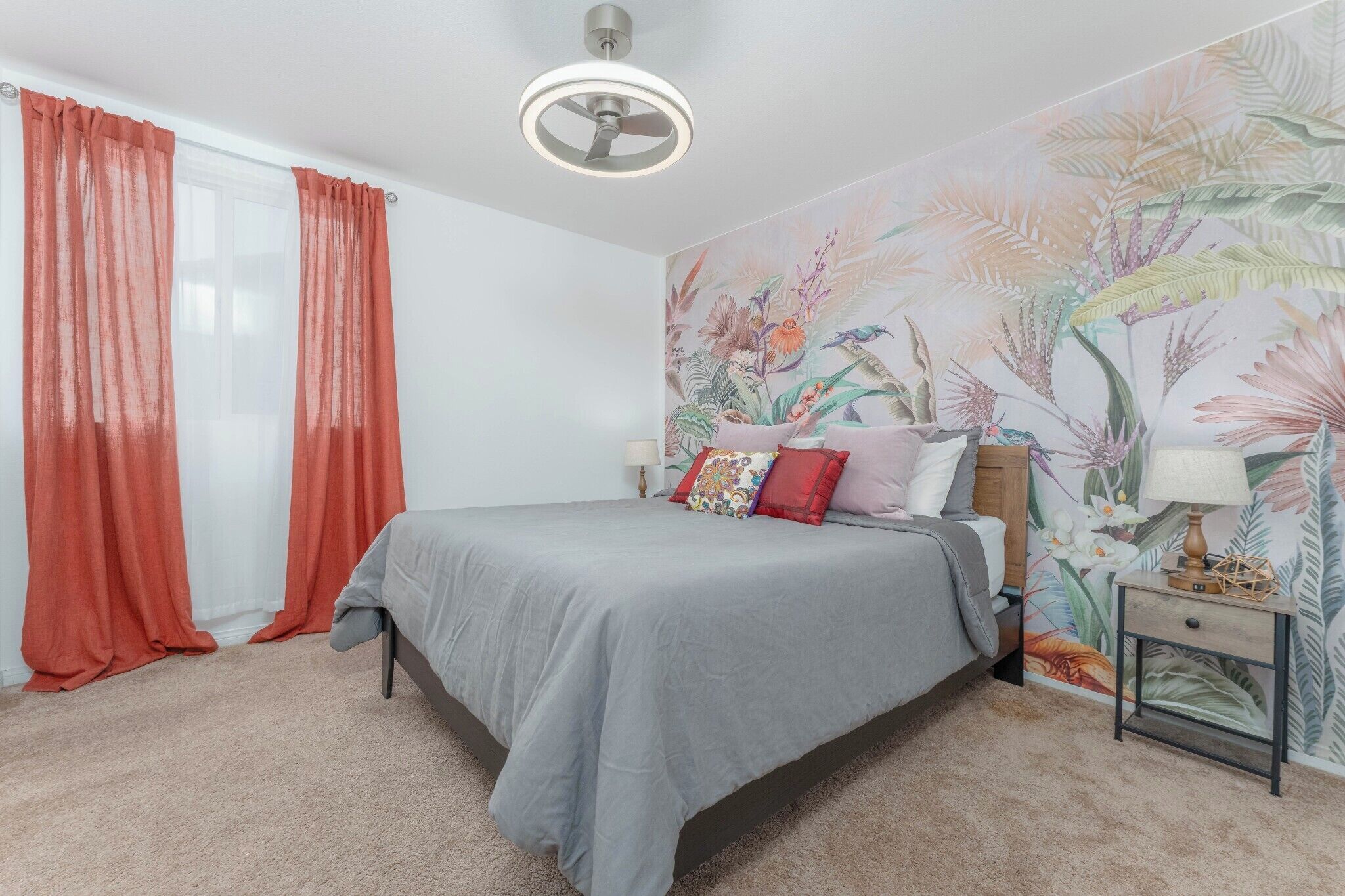 Une chambre à coucher avec un lit, des rideaux, une fresque murale, un ventilateur de plafond et une table de chevet.