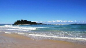 A beach with clear blue water, a sandy shore, and a rocky outcrop in the distance.