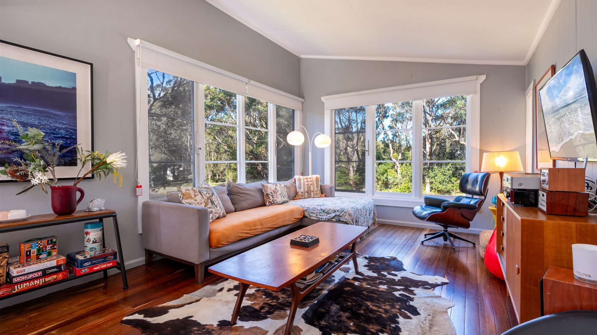 A modern living room with a grey sofa, wooden coffee table, and a cowhide rug. There are large windows with white frames, a bookshelf with board games, and a lamp on a wooden side table.