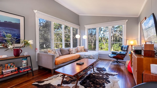 A modern living room with a grey sofa, wooden coffee table, and a cowhide rug. There are large windows with white frames, a bookshelf with board games, and a lamp on a wooden side table.