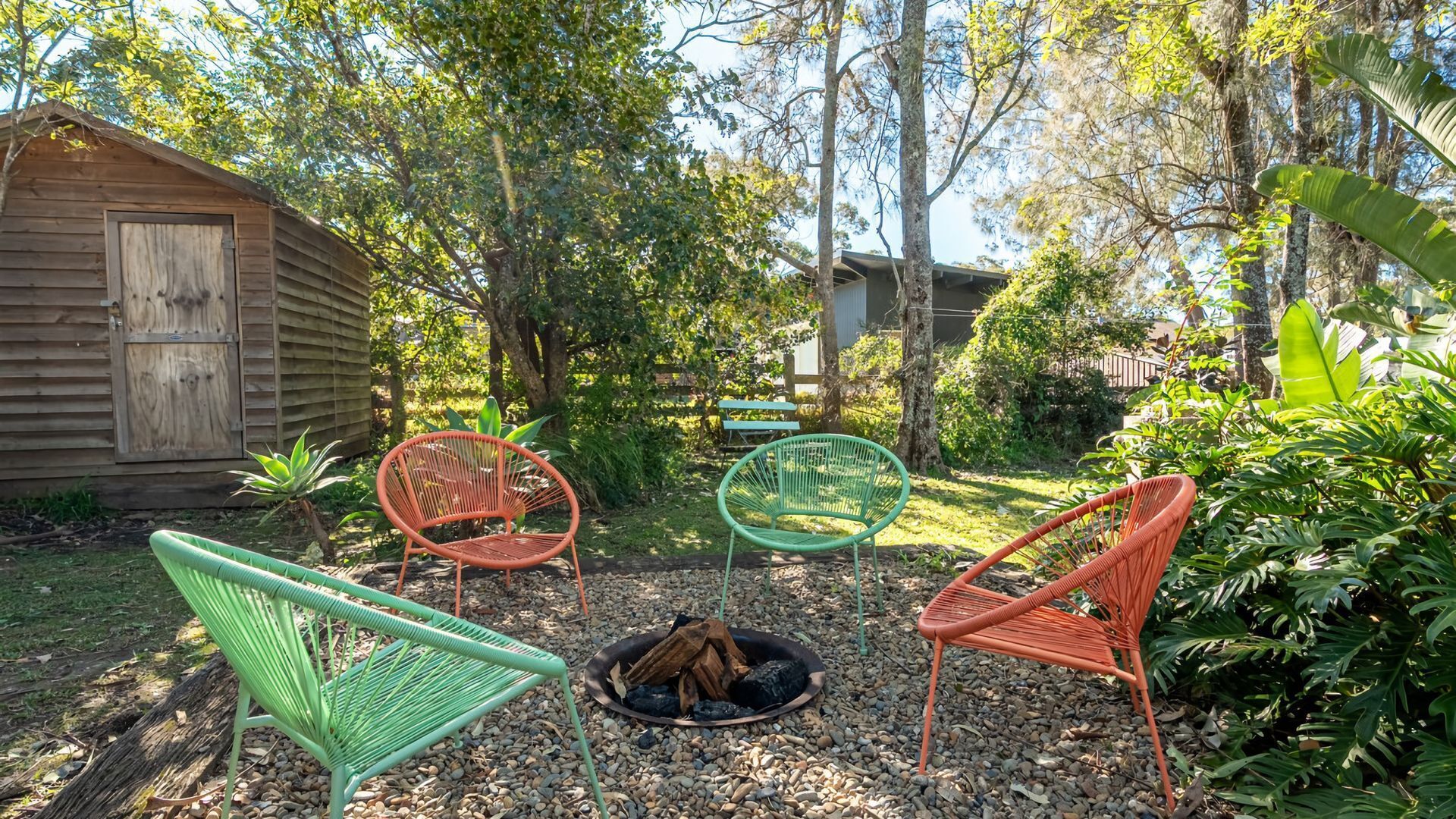 Ein Garten mit einer Holzhütte, einer Feuerstelle und drei Korbstühlen.