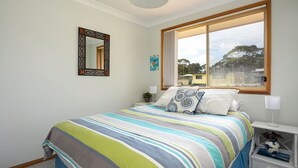 Ein Schlafzimmer mit großem Fenster, einem Bett mit gestreifter Bettwäsche, einem Nachttisch mit Lampe und einem an der Wand befestigten Spiegel.