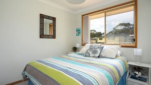 A bedroom with a large window, a bed with striped bedding, a nightstand with a lamp, and a wall-mounted mirror.