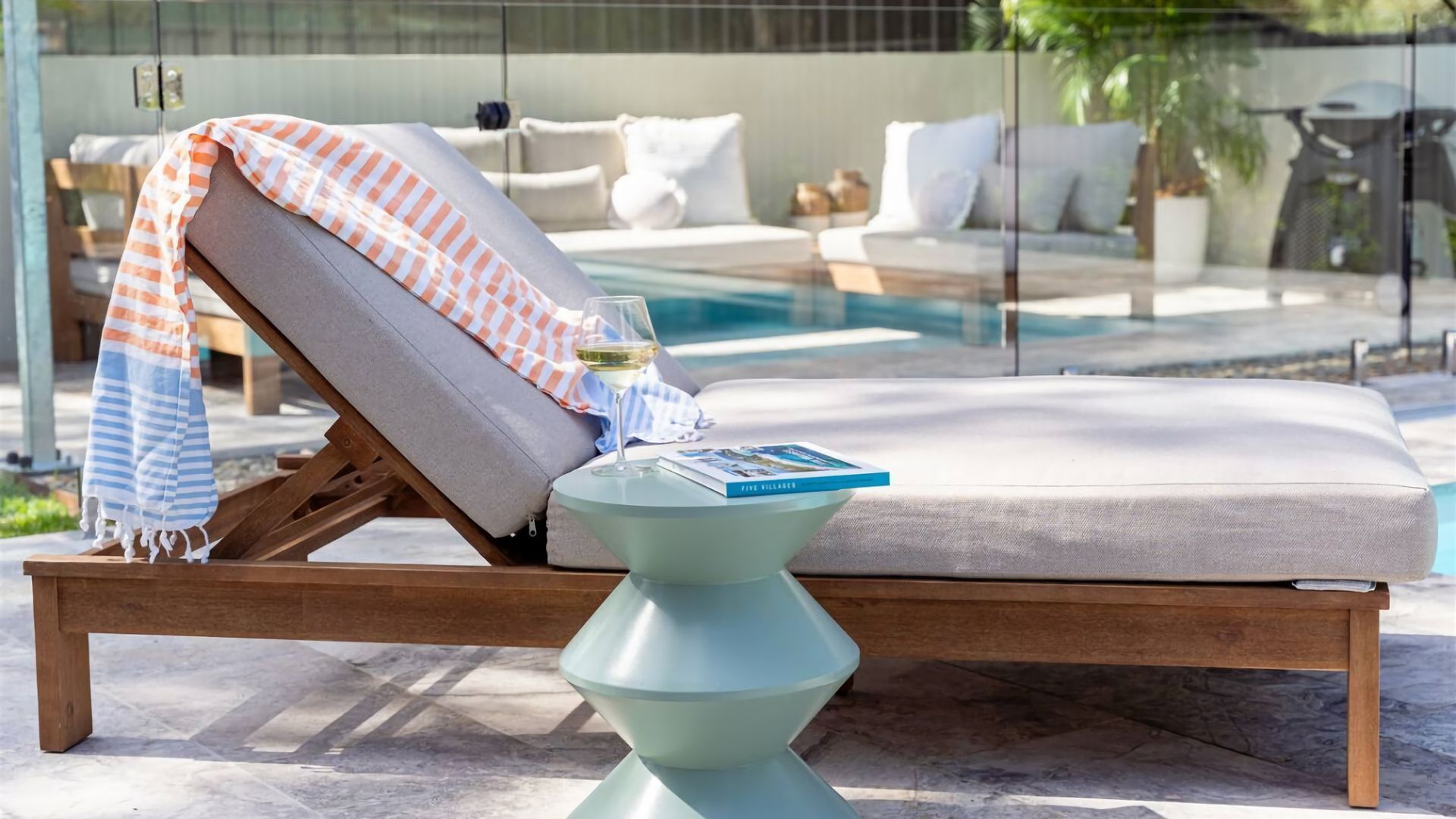 A sun lounger with a striped towel and a glass of white wine, a book titled "FIVE VILLAGES" on a pedestal table, and a pool in the background.