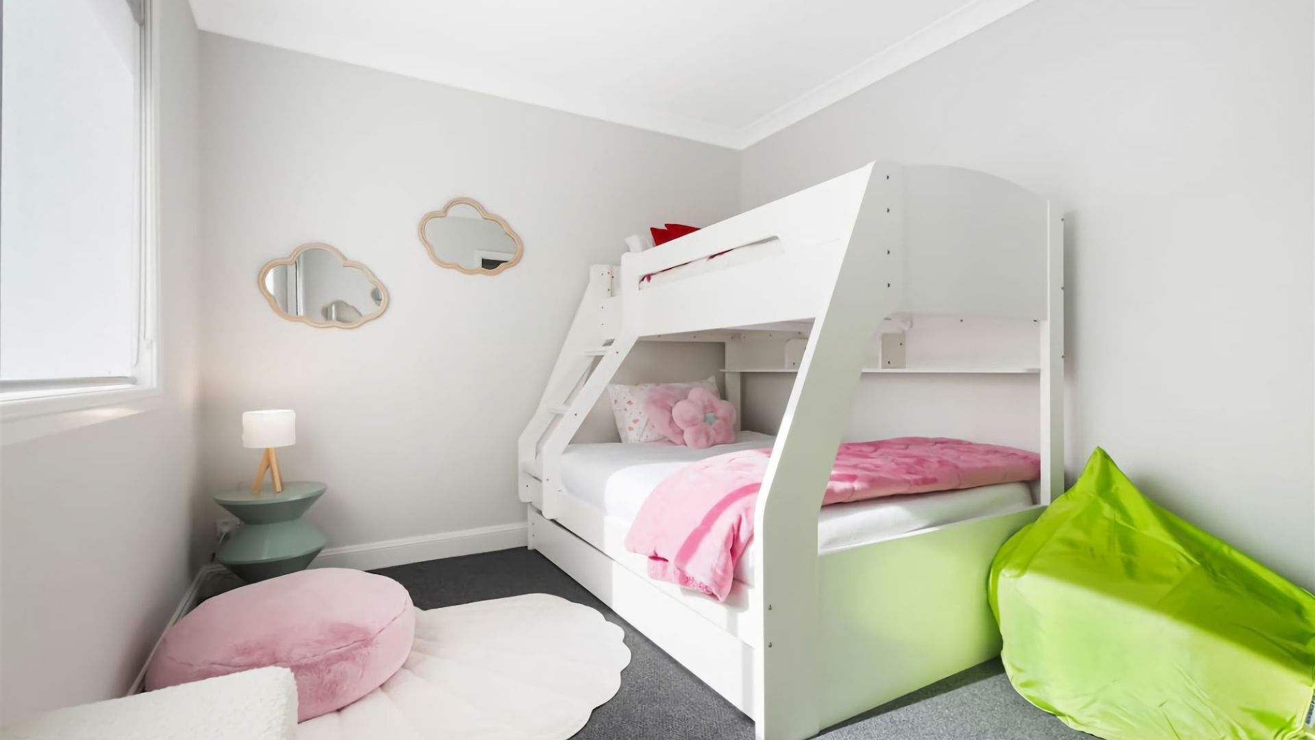 A child's bunk bed with pink bedding, a small bedside table with a lamp, and a green pillow on the floor.