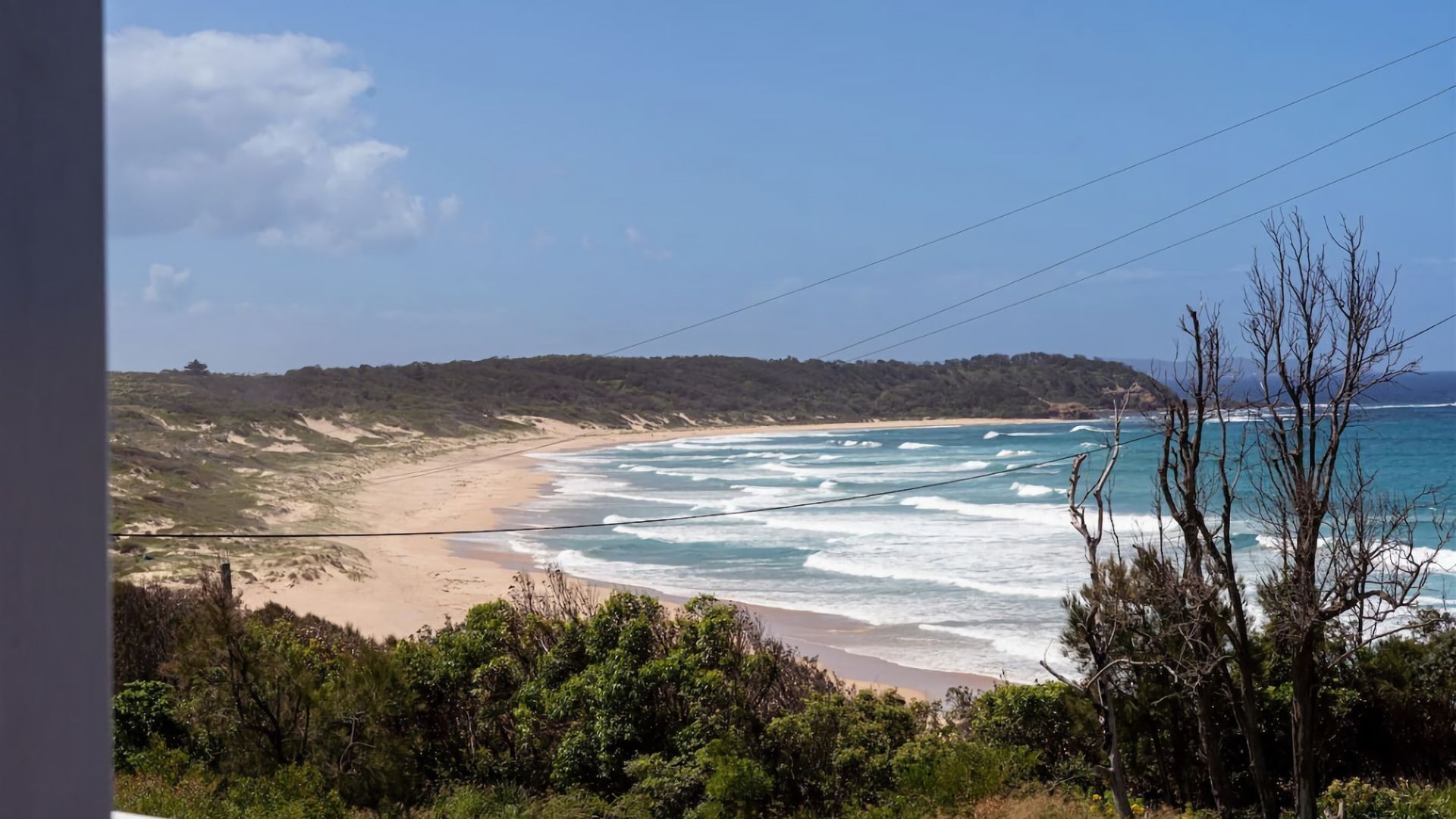 Un paisaje costero con una playa arenosa, olas oceánicas y una colina boscosa al fondo.