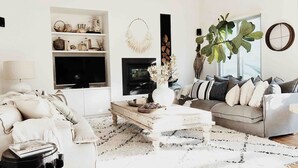 Un salon moderne avec un canapé blanc, une table basse et un téléviseur. On y trouve une cheminée, une grande plante et une bibliothèque.
