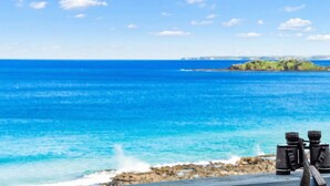 A pair of binoculars on a wooden deck overlooking a clear blue sea with rocky shorelines and distant land.