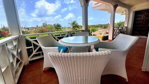 A covered outdoor seating area with wicker furniture and a glass table, overlooking a garden and a clear sky. - Bow Green Villa in Redezvous Bay Steps From Rendezvous Beach (West End)