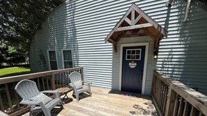Une terrasse avec deux chaises Adirondack et une petite table, une porte bleue ornée d’une pancarte de bienvenue et une rambarde en bois.