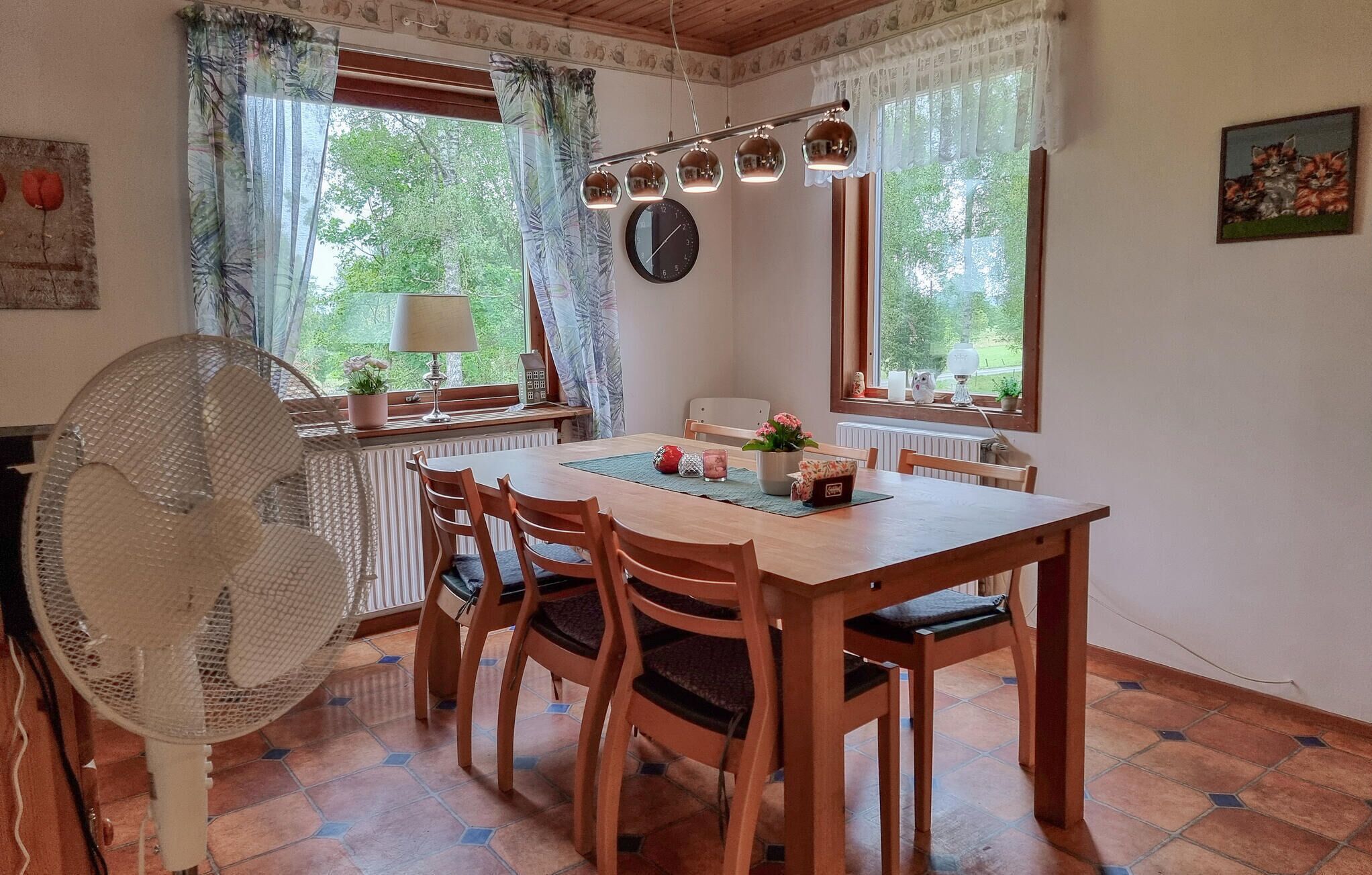 A dining area with a wooden table, four chairs, a fan, a clock, and a window with curtains.