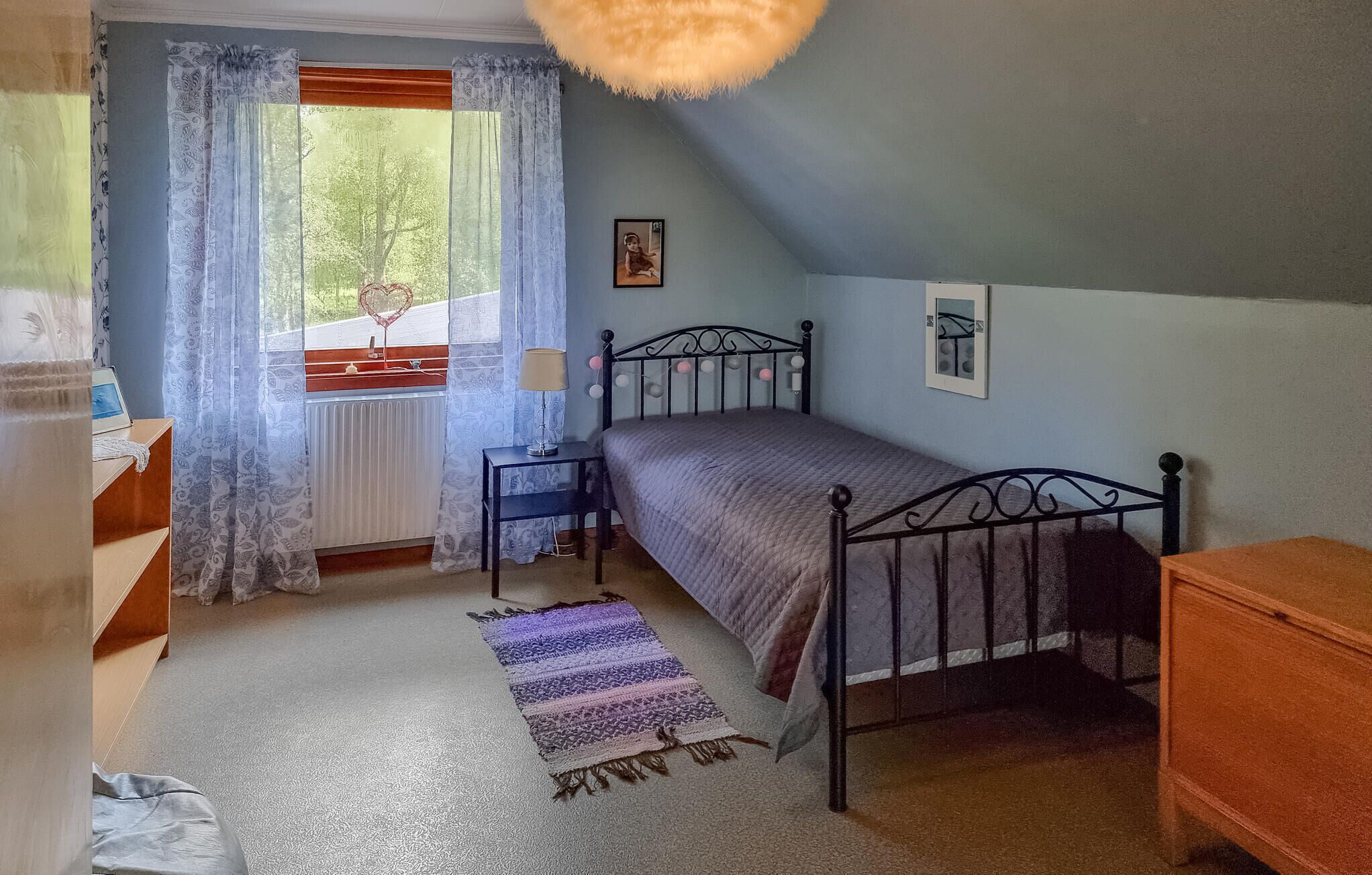Une chambre à coucher comprenant un lit, une table de chevet, une lampe, une commode, une fenêtre avec des rideaux et un radiateur.
