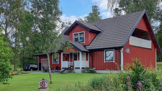 Une maison en bois rouge, avec un toit en pente, un porche et un petit jardin agrémenté d’une nichoir.