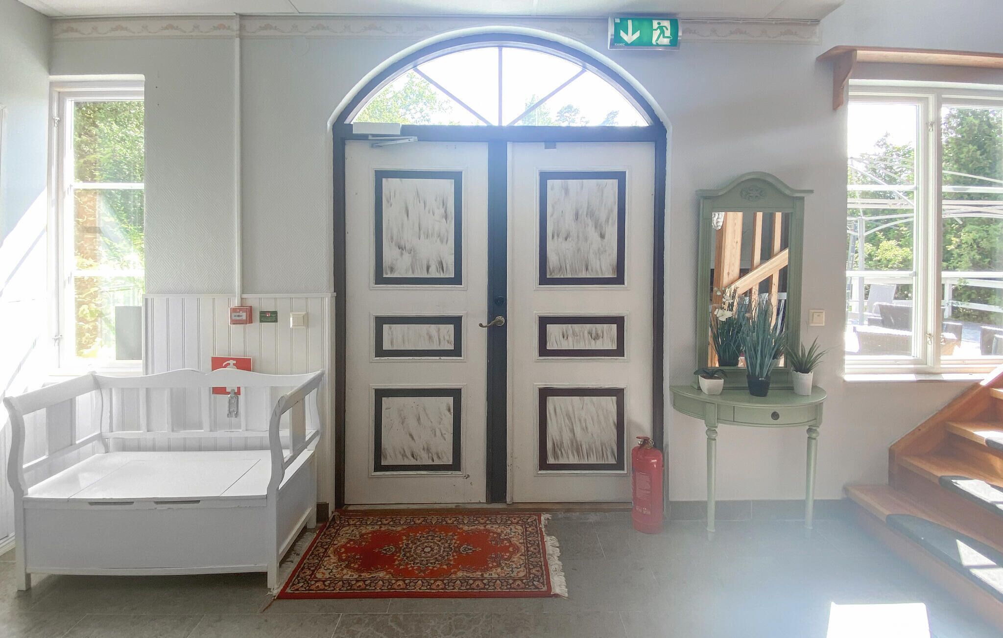 A room with a white bed, a red rug, a green console table with plants, and a large arched window.