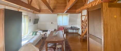 A spacious room with a wooden ceiling, a dining table with chairs, a sofa, and a television mounted on the wall.