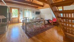 Una habitación amplia con una mesa de comedor de madera, sillas y una butaca roja. Hay un televisor en la pared y una ventana con cortinas.