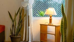 A corner with a standing fan, a potted plant, a wooden nightstand with a lamp, and a window with a view of greenery. - Casa Geranios Panajachel (Panajachel)