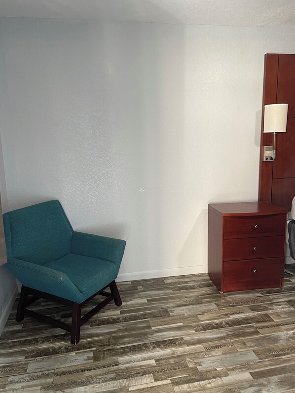 A room with a teal armchair, a wooden dresser with drawers, and a wall-mounted lamp.