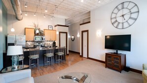 A modern living room with a glass coffee table, a TV, and a kitchen area with a dining set. - Downtown Holland Loft  The "Erie"| 1BR, Walking Distance to Breweries & Dining (Holland)