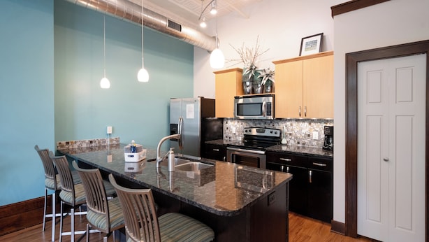 A modern kitchen with a central island, bar stools, stainless steel appliances, and a tiled backsplash. - Downtown Holland Loft  The "Erie"| 1BR, Walking Distance to Breweries & Dining (Holland)