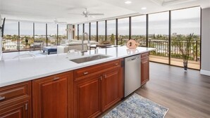 A spacious kitchen with a large island, wooden cabinets, and a dishwasher. It features a balcony with a view of the city and a large window.
