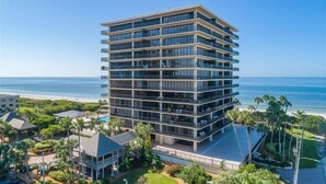 A multi-story hotel with balconies, located near a beach with palm trees and a clear sky.
