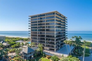 A multi-story hotel with balconies, located near a beach with palm trees and a clear sky.