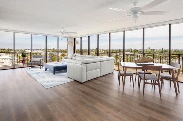 A spacious living room with a white sofa, a dining table with chairs, and a balcony with a view of the city.