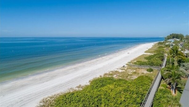 A beach with a clear view of the ocean, a sandy shore, and a pathway leading to a building with greenery and palm trees.