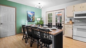 A dining area with a wooden table and black chairs, a white oven, and a green wall with a painting.