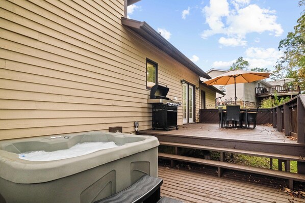 A hot tub on a wooden deck, a barbecue grill, and outdoor furniture under a large umbrella.