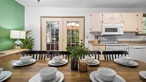 A dining area with a wooden table set for four, surrounded by chairs, with a view of a green wall and a door leading outside.