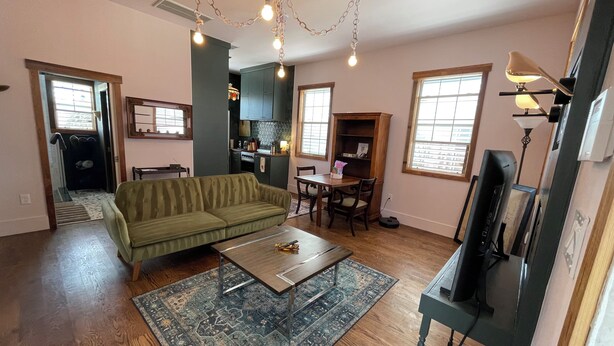 A living room with a green sofa, wooden coffee table, and a patterned rug. There is a kitchen in the background with cabinets and a dining area.
