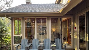 A covered deck with Adirondack chairs and a table, overlooking a garden and a building interior. - Family Friendly Fremont Crossing Home, 3 Kings, Bunk Room, Hot Tub, SHARC Passes (Sunriver)