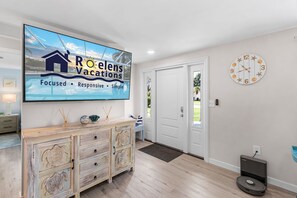 A modern living room with a TV displaying 'Roelens Vacations' branding, a wooden cabinet with drawers, a clock, and a vacuum cleaner.